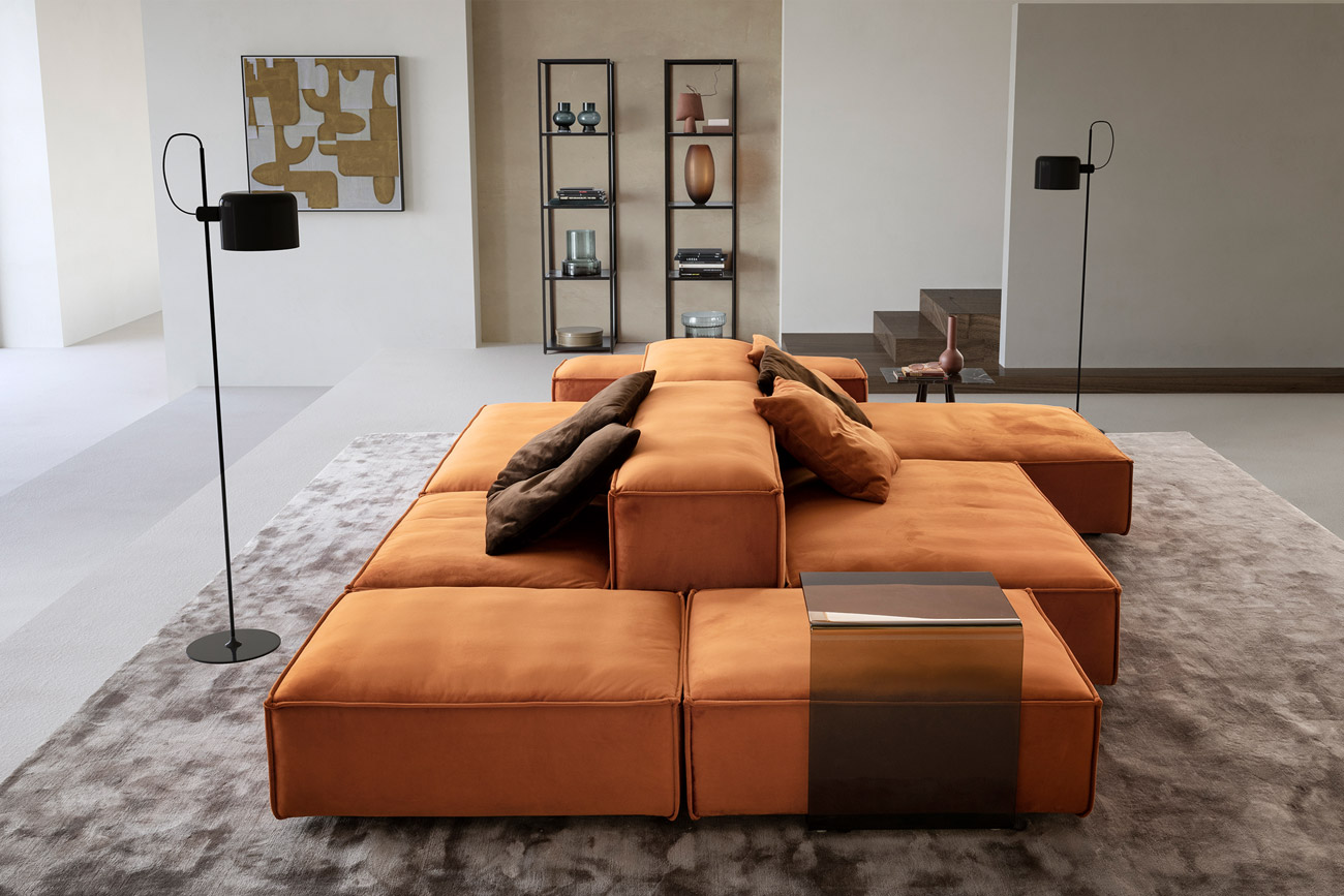 Wohnambiente mit einer Sofakombination des Modells Puzzle mit orangem Stoffbezug, Wurfkissen, C-Table, Four Leg, Tower und Teppich Sense